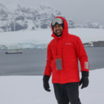 Ateet Ahuja, founder of Complete Getaways, in Antarctica wearing a red Silversea expedition parka with the Silver Cloud expedition ship anchored in the icy waters behind him.