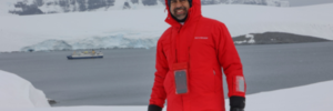 Ateet Ahuja, founder of Complete Getaways, in Antarctica wearing a red Silversea expedition parka with the Silver Cloud expedition ship anchored in the icy waters behind him.