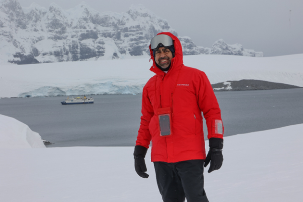 Ateet Ahuja, founder of Complete Getaways, in Antarctica wearing a red Silversea expedition parka with the Silver Cloud expedition ship anchored in the icy waters behind him.