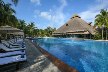 Main pool at El Dorado Royale Riviera Maya
surrounded by tropical landscaping, lounge chairs, and a thatched-roof building
in Riviera Maya, Mexico.